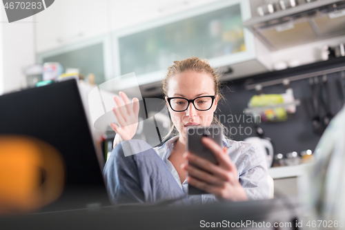 Image of Stay at home and social distancing. Woman in her casual home clothing working remotly from kitchen dining table. Video chatting using social media with friend, family, business clients or partners