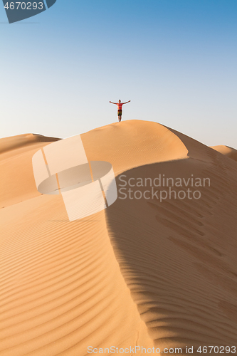 Image of Male traveler standing on the top of dune, arms up to the sky, while traveling sand dessert in Oman.