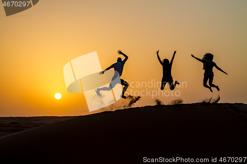 Image of Silhouette of happy traveling people jumping on sand dune and throwing sand in the air in golden sunset hour