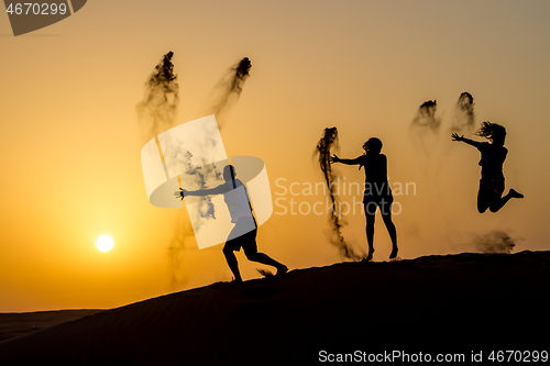 Image of Silhouette of happy traveling people jumping on sand dune and throwing sand in the air in golden sunset hour