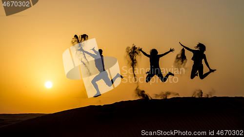 Image of Silhouette of happy traveling people jumping on sand dune and throwing sand in the air in golden sunset hour