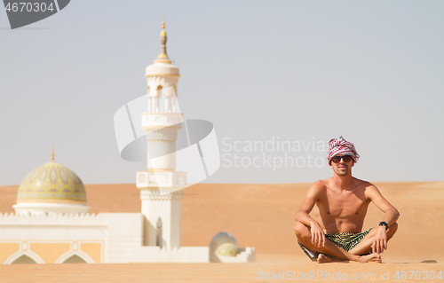 Image of Man traveling in Oman, topless wearing a turban and shorts, sitting on send dune in fron of mosque in desert Oman