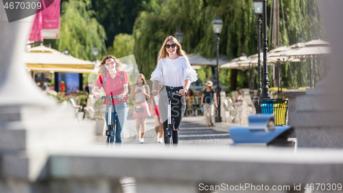 Image of Trendy fashinable teenager girls riding public rental electric scooters in urban city environment. New eco-friendly modern public city transport in Ljubljana, Slovenia