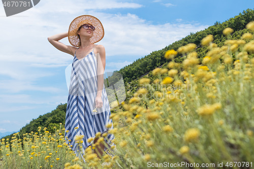 Image of Young woman wearing striped summer dress and straw hat standing in super bloom of wildflowers, relaxing while enjoing beautiful nature of of Adriatic sea coastal nature of Croatia.