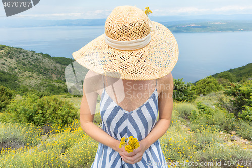 Image of Young woman wearing striped summer dress and straw hat standing in super bloom of wildflowers, holding bouquet of yellow flowers in beautiful nature of Adriatic sea coastal nature of Croatia