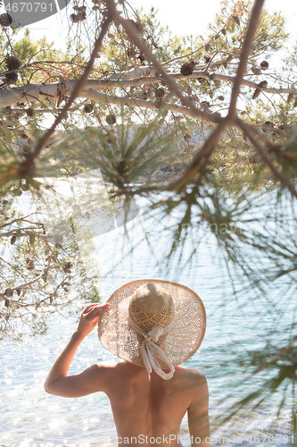 Image of Rear view of topless beautiful woman wearing nothing but straw sun hat realaxing on wild coast of Adriatic sea on a beach in shade of pine tree.