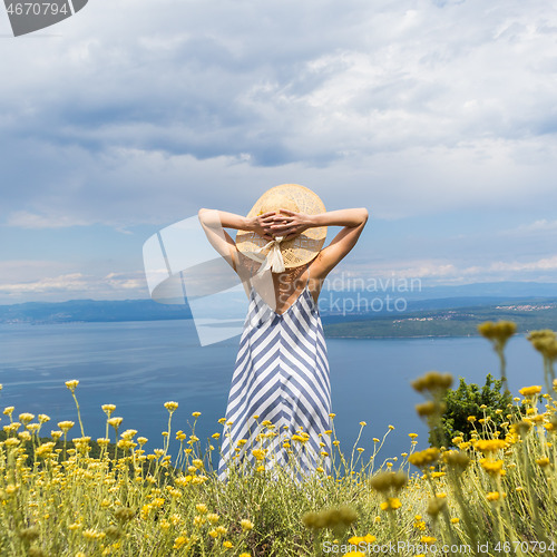 Image of Rear view of young woman wearing striped summer dress and straw hat standing in super bloom of wildflowers, relaxing while enjoing beautiful view of Adriatic sea nature, Croatia