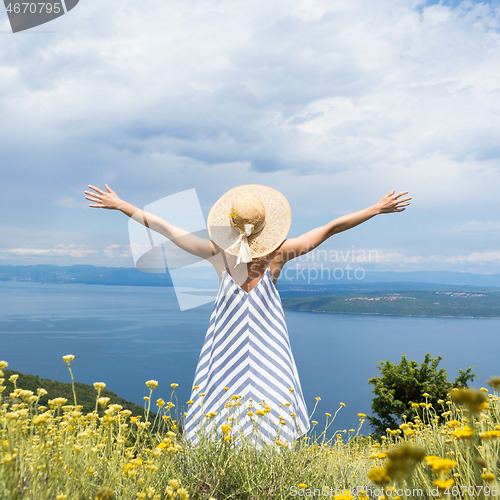 Image of Rear view of young woman wearing striped summer dress and straw hat standing in super bloom of wildflowers, relaxing with hands up to the sky, enjoing beautiful view of Adriatic sea nature, Croatia