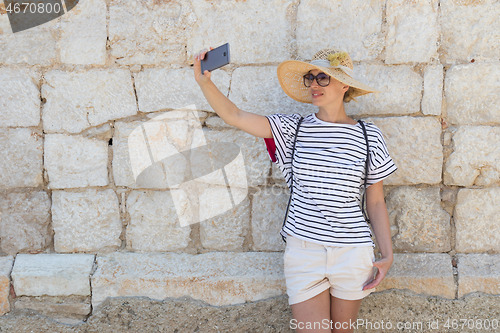 Image of Beautiful young female tourist woman wearing big straw hat, taking self portrait selfie, standing in front of old textured stone wall at old Mediterranean town