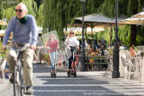 Image of Trendy fashinable teenager girls riding public rental electric scooters in urban city environment. New eco-friendly modern public city transport in Ljubljana, Slovenia