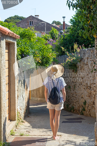 Image of Rear view of beautiful blonde young female traveler wearing straw sun hat sightseeing and enjoying summer vacation in an old traditional costal town at Adriatic cost, Croatia