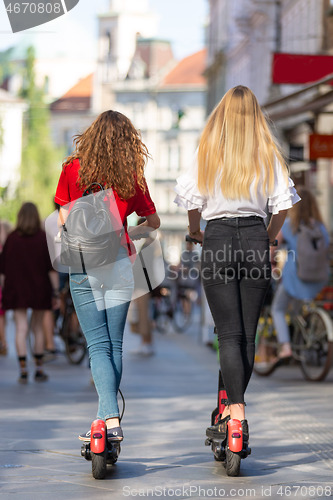 Image of Rear view of trendy fashinable teenager girls riding public rental electric scooters in urban city environment. New eco-friendly modern public city transport in Ljubljana, Slovenia