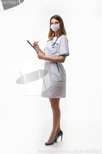 Image of Slender nurse in medical dress and medical mask records data on white background