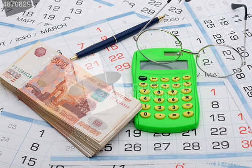 Image of A bundle of money, calculator, glasses and a ballpoint pen lies on the background from the calendar