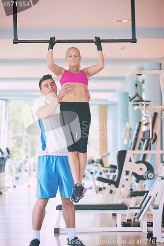 Image of trainer support young woman while lifting on bar in fitness gym