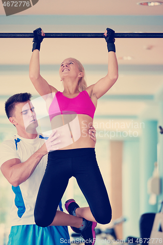 Image of trainer support young woman while lifting on bar in fitness gym