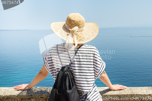 Image of Rear view of woman traveler wearing straw summer hat and backpack,leaning against a stone wall looking at big blue sea and islands in on the horizon.