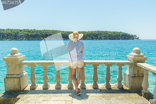 Image of Rear view of woman wearing straw summer hat ,leaning against elegant old stone fence of coastal villa, relaxing while looking at blue Adriatic sea, on Losinj island Croatia.