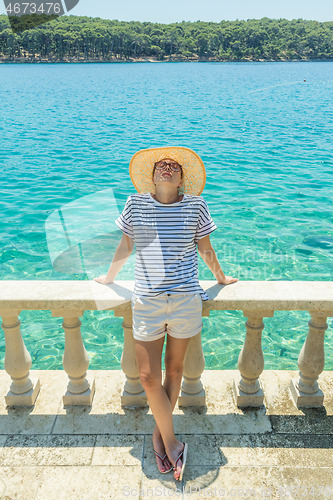 Image of Rear view of woman wearing straw summer hat ,leaning against elegant old stone fence of coastal villa, relaxing while looking at blue Adriatic sea, on Losinj island Croatia.