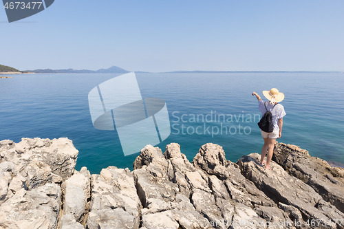 Image of Woman traveler wearing straw summer hat and backpack, standing at edge of the rocky cliff looking and pointing at big blue sea and islands in on the horizon