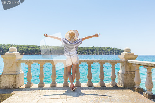 Image of Rear view of happy woman on vacation, wearing straw summer hat ,standing on luxury elegant old stone balcony of coastal villa, relaxing, arms rised to the sun, looking at blue Adriatic sea