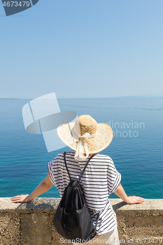 Image of Rear view of woman traveler wearing straw summer hat and backpack,leaning against a stone wall looking at big blue sea and islands in on the horizon.