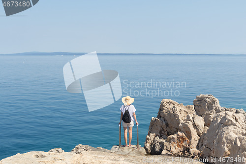 Image of Woman traveler wearing straw summer hat and backpack, standing at edge of the rocky cliff looking at big blue sea and islands in on the horizon