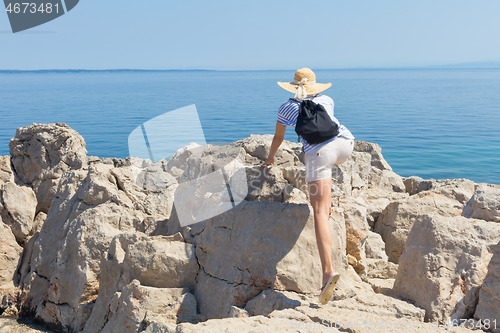 Image of Woman traveler wearing straw summer hat and backpack, climbing at edge of the rocky cliff and looking at big blue sea and islands in on the horizon