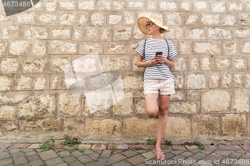 Image of Beautiful young female tourist woman standing in front of old textured stone wall at old Mediterranean town, smiling, holding, smart phone to network on vacationes. Copy space