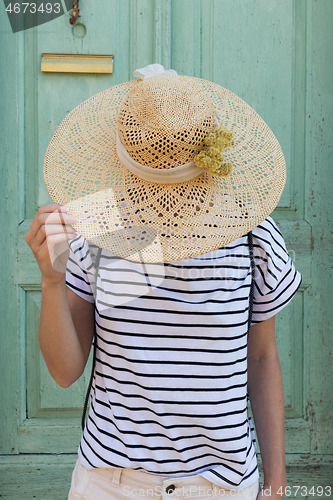 Image of Unrecognizable female tourist woman wearing big straw standing in front of vinatage turquoise wooden door at old Mediterranean town