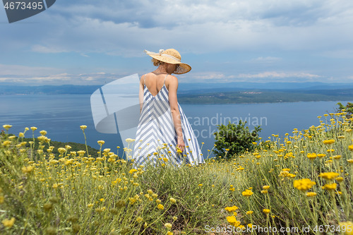 Image of Rear view of young woman wearing striped summer dress and straw hat standing in super bloom of wildflowers, relaxing while enjoing beautiful view of Adriatic sea nature, Croatia