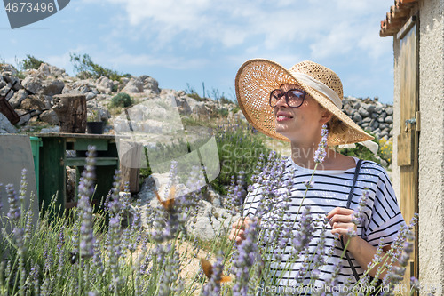 Image of Beautiful blonde young female traveler wearing straw sun hat enjoying summer on Mediterranean cost strolling among lavander flowers on traditional costal village garden