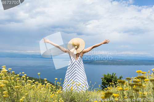 Image of Rear view of young woman wearing striped summer dress and straw hat standing in super bloom of wildflowers, relaxing with hands up to the sky, enjoing beautiful view of Adriatic sea nature, Croatia