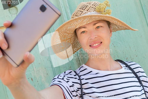 Image of Beautiful young female tourist woman wearing big straw hat, taking self portrait selfie, standing in front of vinatage turquoise wooden door at old Mediterranean town.