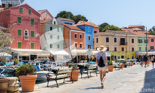 Image of Rear view of beautiful blonde young female traveler wearing straw sun hat sightseeing and enjoying summer vacation in an old traditional costal town of Veli Losinj, Adriatic cost, Croatia