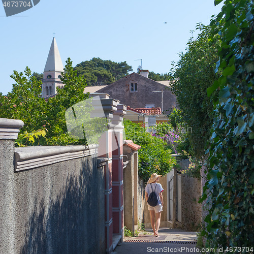 Image of Rear view of beautiful blonde young female traveler wearing straw sun hat sightseeing and enjoying summer vacation in an old traditional costal town of Veli Losinj, Adriatic cost, Croatia