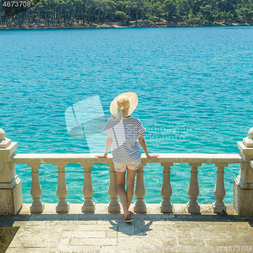 Image of Rear view of woman wearing straw summer hat ,leaning against elegant old stone fence of coastal villa, relaxing while looking at blue Adriatic sea, on Losinj island Croatia.