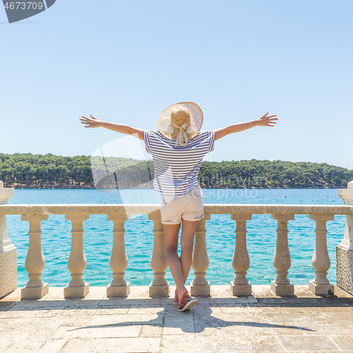 Image of Rear view of happy woman on vacation, wearing straw summer hat ,standing on luxury elegant old stone balcony of coastal villa, relaxing, arms rised to the sun, looking at blue Adriatic sea