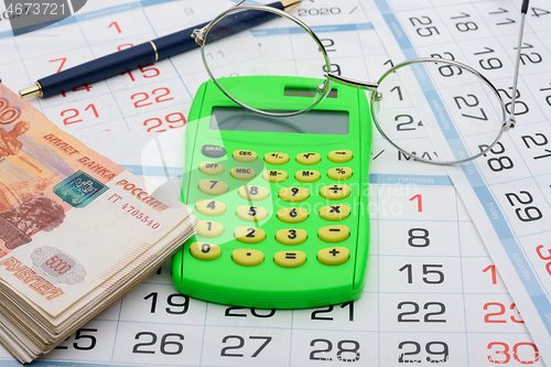 Image of Calculator in the center of the frame, money on the sides, a pen and glasses