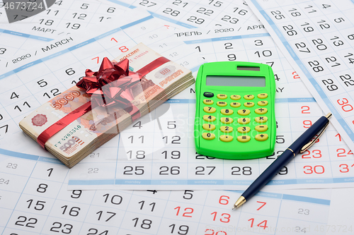 Image of On the background from the calendar lies a bundle of money with a red bow, a calculator and a pen