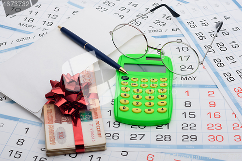 Image of They put a wad of money in an envelope, rewound with a red ribbon and a bow, next to it lies a calculator, a pen, glasses and a background from calendars