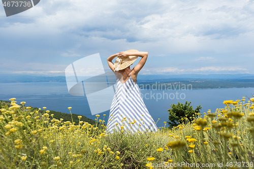 Image of Rear view of young woman wearing striped summer dress and straw hat standing in super bloom of wildflowers, relaxing while enjoing beautiful view of Adriatic sea nature, Croatia