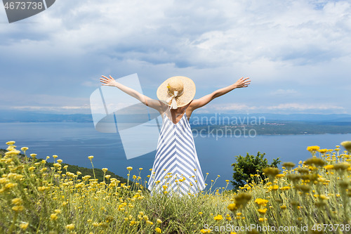 Image of Rear view of young woman wearing striped summer dress and straw hat standing in super bloom of wildflowers, relaxing with hands up to the sky, enjoing beautiful view of Adriatic sea nature, Croatia