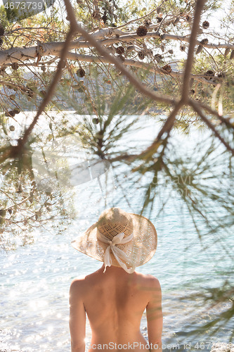 Image of Rear view of topless beautiful woman wearing nothing but straw sun hat realaxing on wild coast of Adriatic sea on a beach in shade of pine tree.