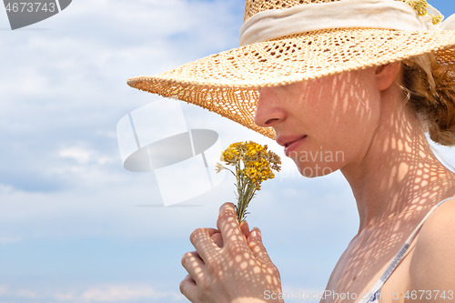 Image of Portrait of young beautiful cheerful woman wearing straw sun hat, smelling small bouquet of yellow wild florets, against blue summer sky