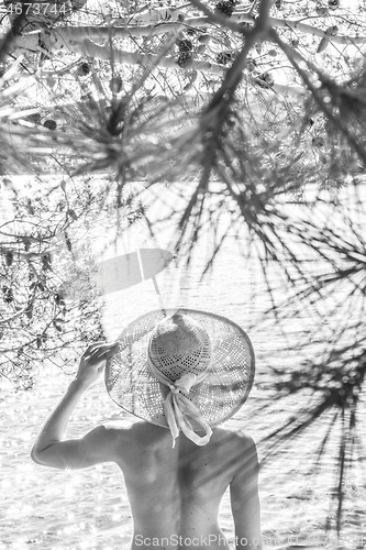 Image of Rear view of topless beautiful woman wearing nothing but straw sun hat realaxing on wild coast of Adriatic sea on beach in shade of pine tree. Relaxed healthy lifestyle concept. Black and white image