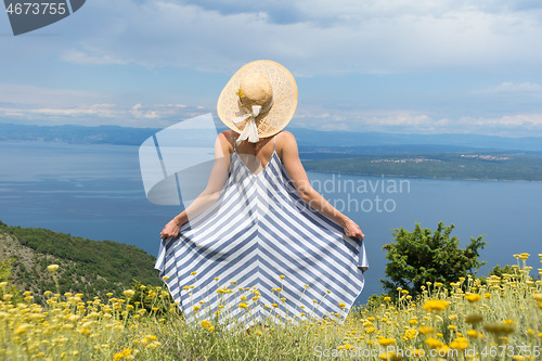 Image of Rear view of young woman wearing striped summer dress and straw hat standing in super bloom of wildflowers, relaxing while enjoing beautiful view of Adriatic sea nature, Croatia