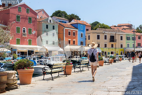 Image of Rear view of beautiful blonde young female traveler wearing straw sun hat sightseeing and enjoying summer vacation in an old traditional costal town of Veli Losinj, Adriatic cost, Croatia