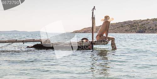 Image of View of unrecognizable woman wearing big summer sun hat tanning topless and relaxing on old wooden pier in remote calm cove of Adriatic sea, Croatia