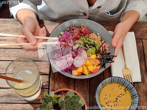 Image of Woman eating tasty colorful healthy natural organic vegetarian Hawaiian poke bowl using asian chopsticks on rustic wooden table. Healthy natural organic eating concept